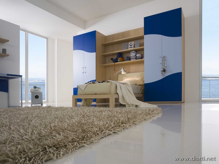 Vista alternativa con particolare sul letto in appoggio al pannello boiserie con mensole; la finitura rovere chiaro si abbina bene al blu ed azzurro dell'armadio.